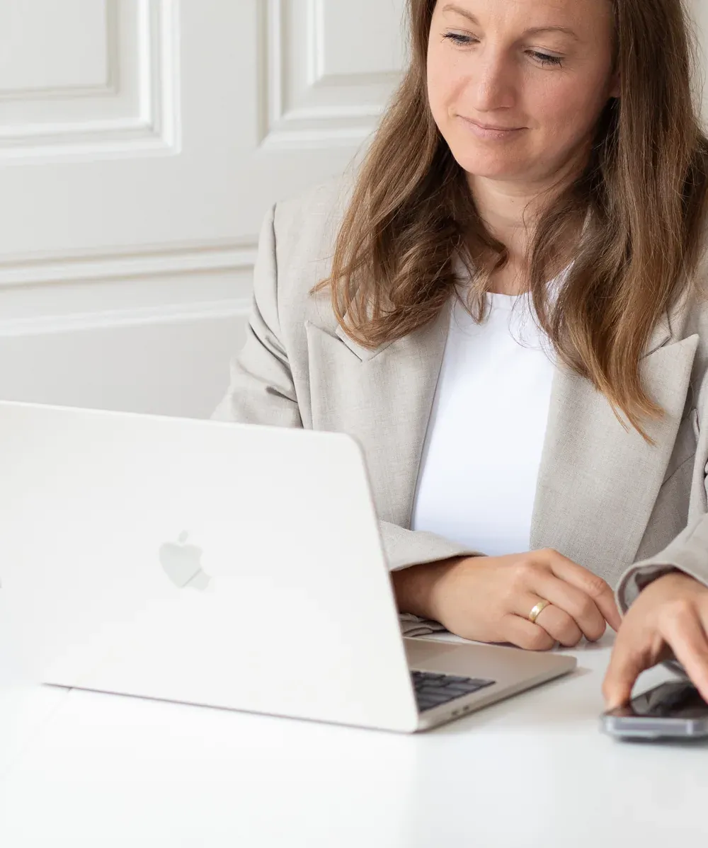 Nadine Fraiss, in a light blazer, sits at a white desk in front of a wall with stucco panelling and looks at her open laptop. One hand rests on the keyboard, the other on a smartphone beside it.